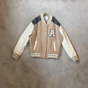 Axel Arigato bomberjacka - Snygg beige bomberjacka med vita ärmar och svarta axelpartier. Jackan har ribbade muddar, två fickor fram och en stor svartvit A-patch på bröstet. Materialmix av tyg och konstläder ger en sportig collegekänsla. Perfekt för dig som gillar streetwear och trendiga detaljer.