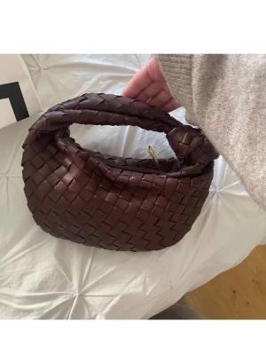 Brun flätad handväska i läder - Säljer en snygg brun handväska i flätad läderimitation med guldfärgad dragkedja. Väskan har en unik knutdetalj på handtaget och en mjuk, rundad form. Perfekt accessoar för att lyfta din outfit.