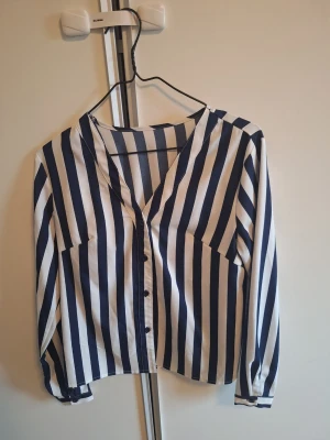 Randig blåvit skjorta med knappar - Snygg skjorta med vertikala blå och vita ränder. Skjortan har v-ringning, långa ärmar och knäppning framtill. Perfekt för dig som gillar en stilren och trendig look.