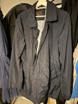 Mörkblå rock från Woolrich - Stilren mörkblå rock från Woolrich med klassisk krage och knappstängning. Rocken har två snedställda fickor framtill och är tillverkad i ett lätt, vattenavvisande material. Perfekt för dig som vill ha en enkel men snygg look.