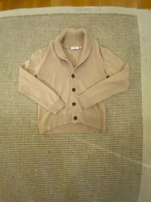 Beige cardigan från RYVLS Stockholm - Snygg beige cardigan från RYVLS Stockholm med stora knappar framtill och bred krage. Koftan har långa ärmar och ribbade muddar vid ärmslut och nederkant. Perfekt för lager på lager och enkel att matcha med olika outfits.