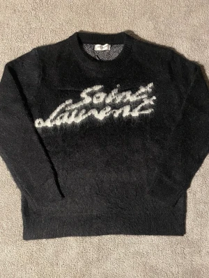 Svart Saint Laurent stickad tröja - Säljer en svart stickad tröja från Saint Laurent med vit logotyp i borstigt garn framtill. Tröjan har rund hals, långa ärmar och ribbade muddar. Materialet ser ut att vara mjukt och fluffigt, troligen mohair eller ullblandning. Perfekt för dig som gillar exklusiva streetwear-plagg.