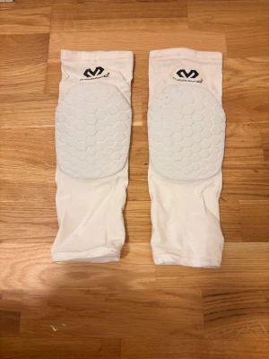 Vita knäskydd från McDavid - Säljer ett par vita knäskydd från McDavid med stötdämpande hexagonmönstrad vaddering på framsidan. Tillverkade i stretchigt material som sitter tight runt benen, perfekta för sporter som basket eller volleyboll. Logga framtill och lång modell som täcker större delen av underbenet.