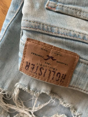 Ljusblå slitna jeans från Hollister - Säljer ett par ljusblå jeans från Hollister med snygga slitningar och hål på benen. Jeansen har klassisk femficksmodell, låg midja och skinny fit. Tillverkade i mjuk denim med stretch för extra komfort. Perfekta för en avslappnad och trendig look.