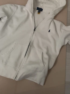 Vit hoodie från Polo Ralph Lauren 14-16 - Säljer en clean vit hoodie från Polo Ralph Lauren med dragkedja och klassisk blå logga på bröstet. Hoodien har huva, kängurufickor och är gjord i mjukt bomullsmaterial. Perfekt för dig som gillar stilrena och bekväma plagg.