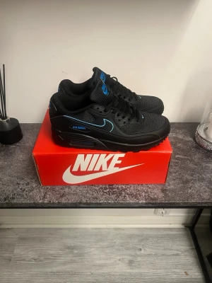 Nike Air Max 90 svart/blå sneakers - Nike Air Max 90 sneakers i svart med blå detaljer och logga. Skorna har en klassisk låg siluett, mesh och läder på ovandelen samt den ikoniska Air Max-sulan för extra komfort. Swoosh-loggan på sidan och blå accenter ger en clean och modern look.