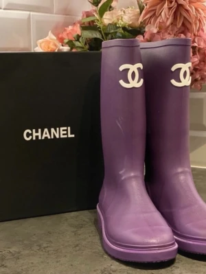Lila gummistövlar från Chanel - Säljer ett par lila höga gummistövlar från Chanel med den ikoniska vita CC-loggan framtill. Stövlarna har en chunky sula och är tillverkade i slitstarkt gummi, perfekta för regniga dagar. Snygg och trendig design som sticker ut.
