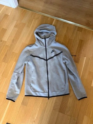 Grå Nike Tech Fleece hoodie - Snygg grå Nike Tech Fleece hoodie med hel dragkedja och svarta detaljer. Har två fickor framtill och en extra ficka med dragkedja på ärmen. Ikonisk Nike-logga på bröstet och skön huva. Perfekt för en sportig och avslappnad stil.