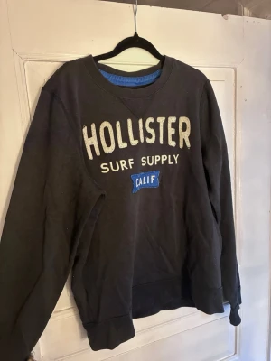 Marinblå/svart sweatshirt från Hollister - Cool marinblå/svart sweatshirt från Hollister med broderad text 'HOLLISTER SURF SUPPLY CALIF' i vitt och blått framtill. Klassisk rund halsringning och ribbade muddar. Perfekt för en avslappnad streetwear-look. Passar snarare M/L