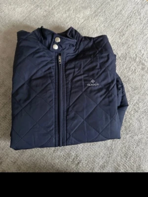 Marinblå quiltad jacka från Gant - Snygg marinblå quiltad jacka från Gant med dragkedja framtill och tryckknappar vid kragen. Jackan har diskret logga på bröstet och rutmönstrad design. Perfekt för dig som gillar stilrena och klassiska plagg.