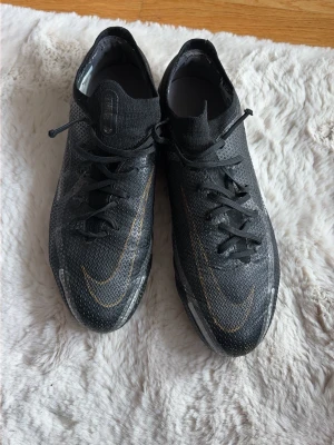 Nike Phantom GT2 Elite FG Svarta Fotbollsskor - Säljer ett par Nike Phantom GT2 Elite fotbollsskor i svart med guldiga detaljer och snörning. Skorna har en strömlinjeformad design och är tillverkade i ett lätt syntetmaterial för bästa bollkontroll på planen. Perfekta för dig som vill ha stilrena och grymma skor till matchen. Använda, lite slitage.
