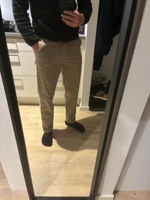 Beige chinos med rak passform - Snygga beige chinos med rak passform och klassiska fickor fram. Byxorna har bälteshällor och stängs med dragkedja och knapp. Perfekta för en clean och stilren look. Materialet känns mjukt och bekvämt, troligen bomull eller bomullsblandning.