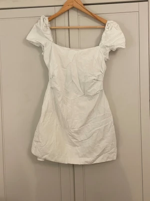 Vit kort klänning med puffärm - Söt vit kort klänning med puffärmar och öppen rygg med dekorativa band. Klänningen har en rak passform och är tillverkad i ett lätt tyg, perfekt för varma dagar. Den har en enkel men trendig design som passar till många olika stilar.