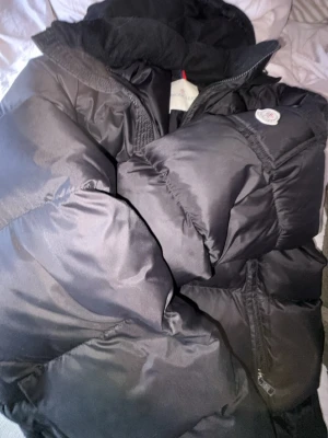 Svart pufferjacka från Moncler - Säljer en svart pufferjacka från Moncler med stor huva och dragkedja framtill. Jackan har en klassisk Moncler-logga på ärmen och är riktigt tjock och varm, perfekt för kalla vinterdagar eller vår dagar. Materialet är glansigt och quiltat för en modern look.