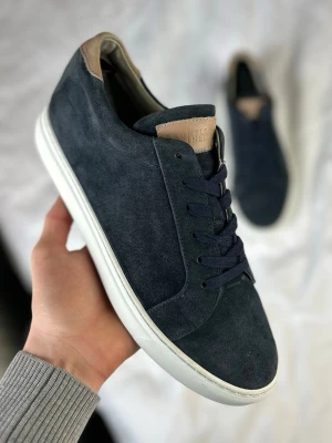 Mörkblå mockasneakers från Brunello Cucinelli - Stilrena sneakers i mörkblå mocka från Brunello Cucinelli med vita sulor och beige detaljer på häl och plös. Klassisk låg modell med matchande mörkblå skosnören. Perfekta för dig som gillar minimalistisk och clean stil.