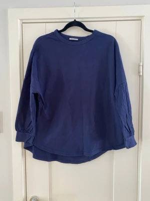 Mörkblå topp med 3/4 ärm - En enkel och stilren mörkblå topp med rund halsringning och 3/4 ärmar. Toppen har en lös passform och breda muddar vid ärmsluten. Perfekt att matcha med jeans eller kjol för en avslappnad look. Tröjan är lite blekt precis på armarna i övrigt i bra begagnat skick.  Mått Över bröstet: ca 70 cm Längd från axeln och ner: ca 63 cm  100% bomull  Kommer från ett djur och rökfritt hem