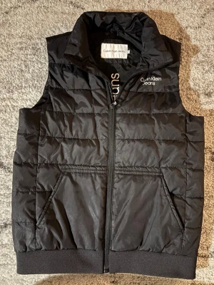 Svart dunväst från Calvin Klein Jeans - Snygg svart dunväst från Calvin Klein Jeans med diskret logga på bröstet. Västen har dragkedja framtill, två fickor och ribbade muddar nedtill. Perfekt för lager på lager och ger en clean streetwear-look.