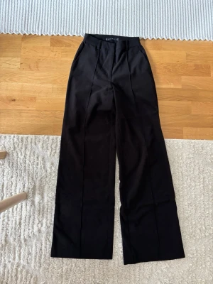 Svarta vida kostymbyxor från Amisu - Säljer ett par svarta kostymbyxor från Amisu med hög midja och vida ben. Byxorna har pressveck framtill, hällor för skärp och sidofickor. Perfekta för en clean och stilren look.