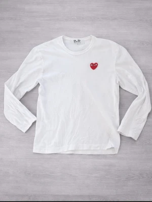 Vit långärmad tröja Comme des Garçons - Vit långärmad tröja från Comme des Garçons PLAY med det ikoniska röda hjärtat med ögon broderat på bröstet. Tillverkad i mjuk bomull och har en klassisk rund halsringning. Perfekt för en clean och stilren look. 3 små håll som kan ses på bilden.