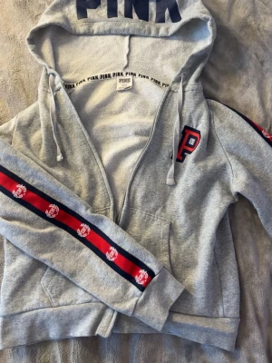 Grå hoodie från PINK Victoria's Secret - Snygg grå hoodie med dragkedja från PINK Victoria's Secret. Stor huva med trycket 'PINK', röda och marinblå ränder med logga längs ärmarna och ett broderat 'P' på bröstet. Mjuk bomullsblandning och kängurufickor framtill. Perfekt för en chill och sportig look.