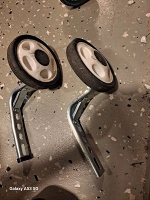 Stödhjul till cykel, metall och plast - Säljer ett par stödhjul till cykel med vita plasthjul och svarta gummikanter. Fästena är i metall med flera hål för justering. Perfekt för att hjälpa till när man lär sig cykla.