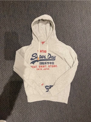 Grå Superdry hoodie med tryck (reserverad) - Snygg ljusgrå hoodie från Superdry med stor logga och text i blått och rött på bröstet. Klassisk huva med dragsko och känguruficka framtill. Tillverkad i mjukt bomullsmaterial, perfekt för en avslappnad streetwear-look.