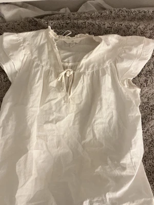 Vit blus med knyt från H&M - Söt vit blus från H&M med korta ärmar och knytband vid halsen. Blusen har en avslappnad passform och lätt rynkade detaljer vid kragen. Tillverkad i ett tunt och luftigt material. Perfekt för en enkel och stilren look.