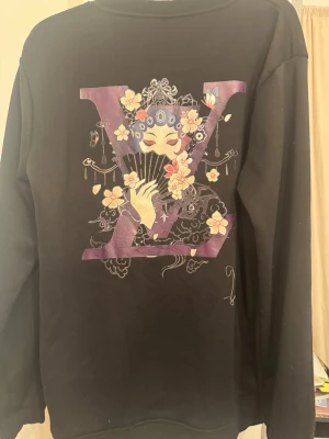 Svart sweatshirt med konstnärligt tryck - Cool svart sweatshirt med stort konstnärligt tryck på ryggen och mindre tryck fram. Motivet har en stor lila bokstav V, blommor och en maskerad figur i asiatiskt inspirerad stil. Tröjan har rund hals och långa ärmar, perfekt för en unik streetwear-look.