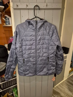 Grå lättviktsjacka med huva - Snygg grå lättviktsjacka med huva och dragkedja framtill. Jackan har två praktiska fickor med dragkedja. Tillverkad i lätt och värmande material.