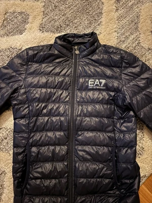 Mörkblå pufferjacka från EA7 Emporio Armani - Mörkblå pufferjacka från EA7. Jackan har dragkedja framtill, två sidofickor med dragkedja och logga på bröstet. Nypris 2199kr