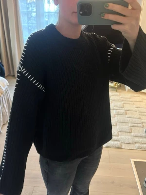 Svart stickad tröja med vita detaljer - Säljer en svart stickad tröja med vita sömdetaljer längs ärmar och axlar. Tröjan har en avslappnad passform, rund halsringning och långa ärmar. Perfekt för dig som gillar en enkel men ändå unik look.