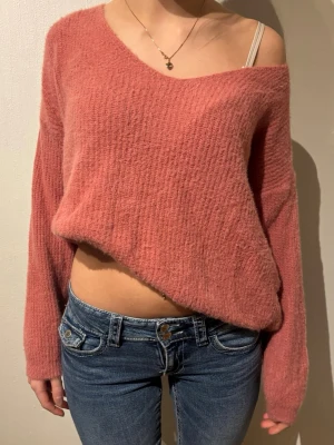 Rosa stickad tröja med v-ringning - Mysig rosa stickad tröja med lös passform och djup v-ringning. Tröjan har långa ärmar och är tillverkad i ett fluffigt, mjukt material som ger en avslappnad look. Perfekt att bära off-shoulder för en trendig stil.