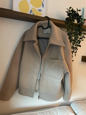 Beige kappa från NA-KD - Stilren beige kappa från NA-KD med dragkedja framtill och stor bröstficka. Kappan har bred krage och raka ärmar, tillverkad i ett tjockare ull-liknande material som ger en trendig och avslappnad look.
