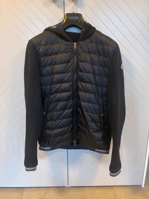 Moncler cardigan  - Snygg svart pufferjacka från Moncler med huva och quiltad front. Ärmarna är i mjukt tyg och har ribbade muddar med vita detaljer. Jackan har dragkedja framtill och Moncler-logga på ärmen. Perfekt för kalla dagar och riktigt stilren. För frågor hör av dig!