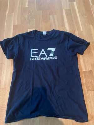 Svart EA7 Emporio Armani t-shirt - Snygg svart t-shirt från EA7 Emporio Armani med stort vitt tryck på bröstet. Klassisk rund hals och korta ärmar. Tillverkad i mjuk bomull för skön känsla och enkel stil.