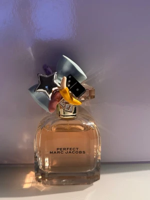 Perfect parfym från Marc Jacobs - En unik parfymflaska från Marc Jacobs, modellen Perfect. Flaskan är transparent med ljusrosa doftvätska och har ett kreativt lock dekorerat med färgglada detaljer som en blå rosett, silverstjärna och banan. Perfekt för dig som gillar lekfull design. 50 ml. Ordinarie pris 1355 kr.