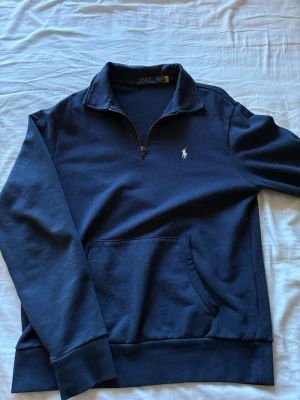 Mörkblå half-zip tröja Polo Ralph Lauren - Mörkblå långärmad tröja från Polo Ralph Lauren med half-zip och klassisk vit broderad logga på bröstet. Tröjan har en känguruficka framtill och ribbade muddar vid ärmslut och nederkant. Perfekt för en avslappnad men stilren look. Knappt använd