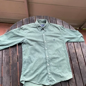 Grön skjorta från Ralph Lauren - Klassisk grön skjorta från Ralph Lauren i 100% bomull. Skjortan har lång ärm, knappar framtill och den ikoniska broderade loggan på bröstet. Perfekt för en stilren och avslappnad look. Skrolla till sista bilden för en fet Ferrari!