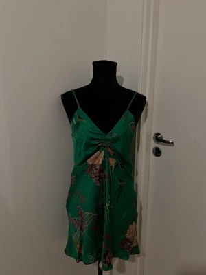 Grön blommig kort klänning i satin - Snygg grön kort klänning med blommigt mönster och tunna axelband. Klänningen har v-ringning och en snygg rynkdetalj framtill. Materialet är glansigt satinliknande tyg som ger en lyxig känsla. Perfekt för sommarens alla tillfällen.