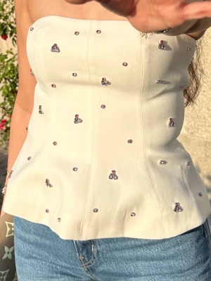 Vit korsett-topp med broderade detaljer - Säljer en snygg vit korsett-topp med broderade små blommor och pärlliknande detaljer. Toppen är axelbandslös och har en figurnära passform som ger en stilren siluett. Perfekt att matcha med jeans eller kjol för en trendig look. Har även en dragkedja 