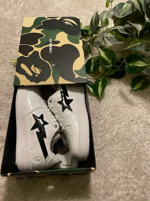 BAPE Skor  - Säljer ett par vita BAPE STA sneakers i blankt läder med den ikoniska svarta stjärnan på sidan. Skorna har klassisk låg siluett och kommer i originalkartong med camomönster. Perfekta för dig som gillar streetwear och vill sticka ut.