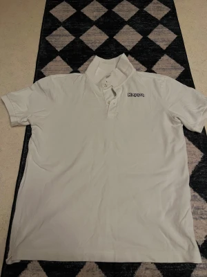 Vit Kappa piké med krage - Klassisk vit pikétröja från Kappa med krage och knappar framtill. Diskret Kappa-logga på bröstet. Perfekt till jeans eller shorts för en clean och sportig look.