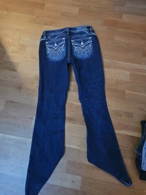 Blå Miss Me jeans med broderi - Säljer ett par blå jeans från Miss Me med snygga broderade bakfickor och vita kontrastsömmar. Jeansen har bootcut-modell och detaljerade ficklock med knappar. Perfekta för dig som vill sticka ut lite extra!