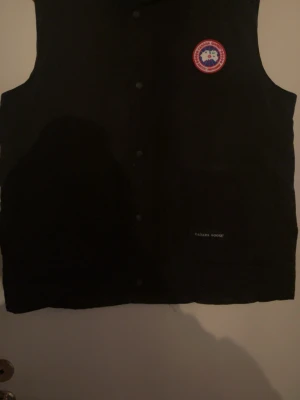 Svart dunväst från Canada Goose - Snygg svart dunväst från Canada Goose med klassisk logga på bröstet och diskret varumärkesbrodyr vid fickan. Västen har tryckknappar framtill och är perfekt för lager-på-lager-stil. Materialet är slitstarkt och håller värmen riktigt bra.