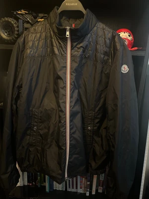 9/10 Svart vindjacka från Moncler  - Snygg svart vindjacka från Moncler med quiltade detaljer upptill och klassisk logotyp på ärmen. Jackan har dragkedja med röd, vit och blå kant, två fickor framtill med knappstängning och hög krage. Tillverkad i lätt, glansigt material som står emot vind.
