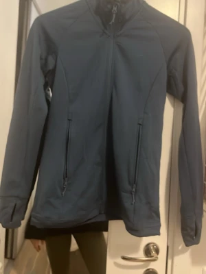 Mörkblå vindjacka från Bergans XS - Sportig mörkblå vindjacka från Bergans med hög krage och dragkedja framtill. Jackan har två praktiska fickor med dragkedja och är tillverkad i ett lätt syntetmaterial. Perfekt för utomhusaktiviteter och vardagsäventyr. Lite fugursyd.