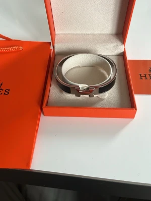 Svart Hermès Clic H armband - OBS. Går inte att låsa men går att bära och den sitter safe. Därav låga pris. Snyggt svart Hermès Clic H armband i emalj och silverfärgad metall. Armbandet har den ikoniska H-detaljen framtill och kommer i originalask med Hermès-logga. Perfekt accessoar för att lyfta din outfit.