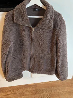 Brun teddyjacka från 157 - Mysig brun teddyjacka från 157 med dragkedja framtill och två stora fickor. Jackan har en hög krage och är gjord i fluffigt teddy-material som ger en avslappnad och trendig look. Perfekt för kyliga dagar och enkel att matcha med olika outfits.