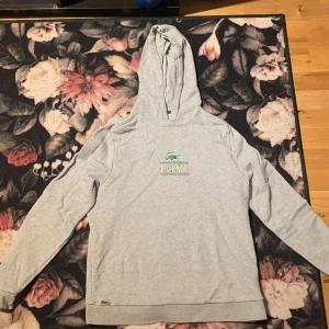 Grå hoodie från Lacoste med logga - Snygg grå hoodie från Lacoste med stor grön logga och krokodil på bröstet. Klassisk passform med huva och långa ärmar. Tillverkad i mjukt bomullsmaterial som känns skönt mot huden. Perfekt för en avslappnad och stilren look.