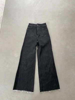 Svarta raka jeans från Zara - Säljer ett par svarta raka jeans från Zara med hög midja och råa, fransiga benslut. Jeansen har stora patchfickor fram och är tillverkade i ett klassiskt denimtyg. Perfekta för en trendig och avslappnad look.